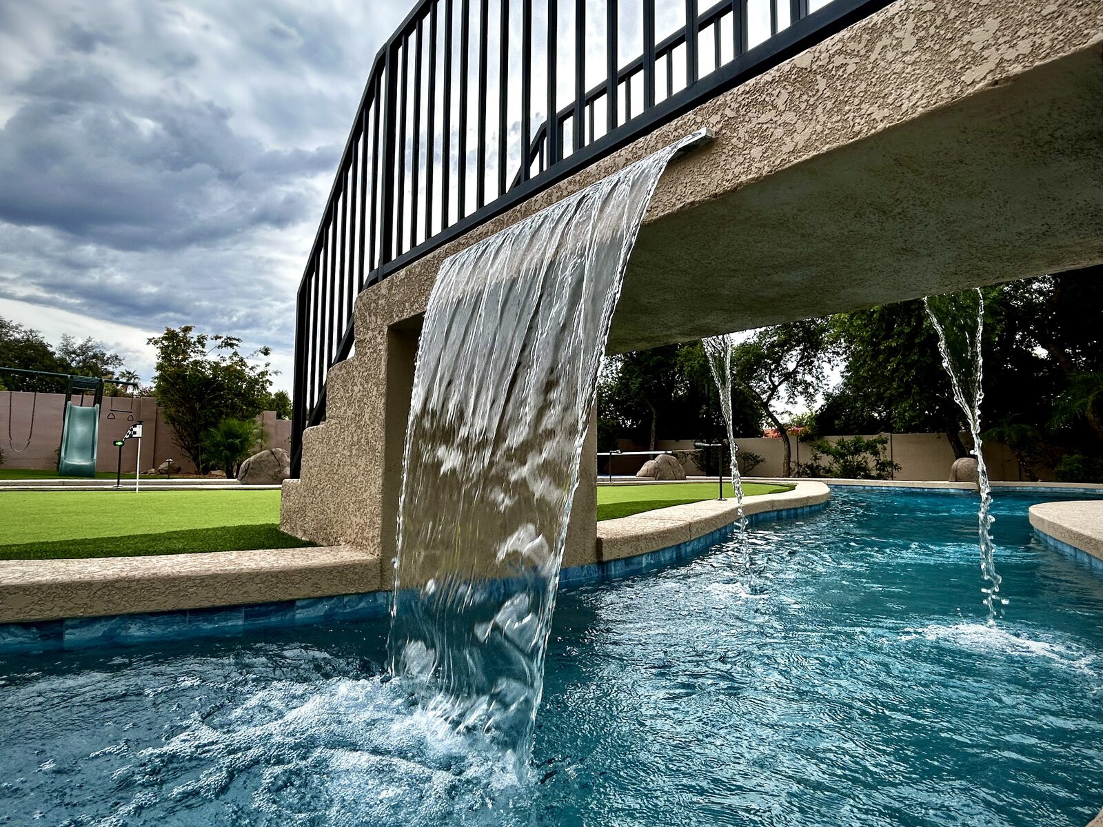Water-level view of the bridge's twin waterfalls against a dramatic cloudy sky, with the putting green and playground beyond