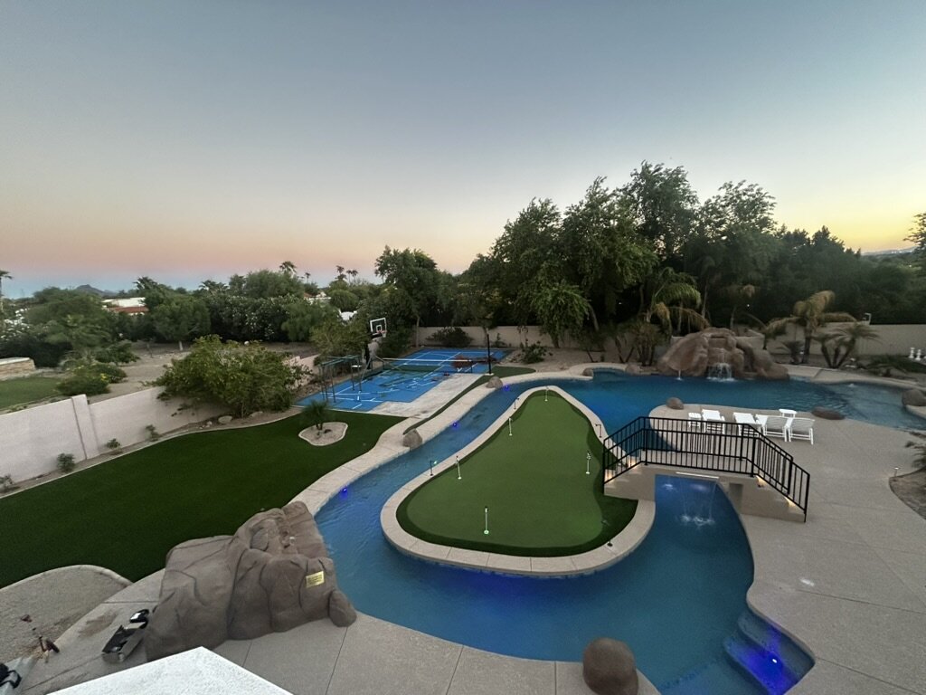 Daytime aerial view of the completed backyard — pool, lazy river, bridge, and putting-green island