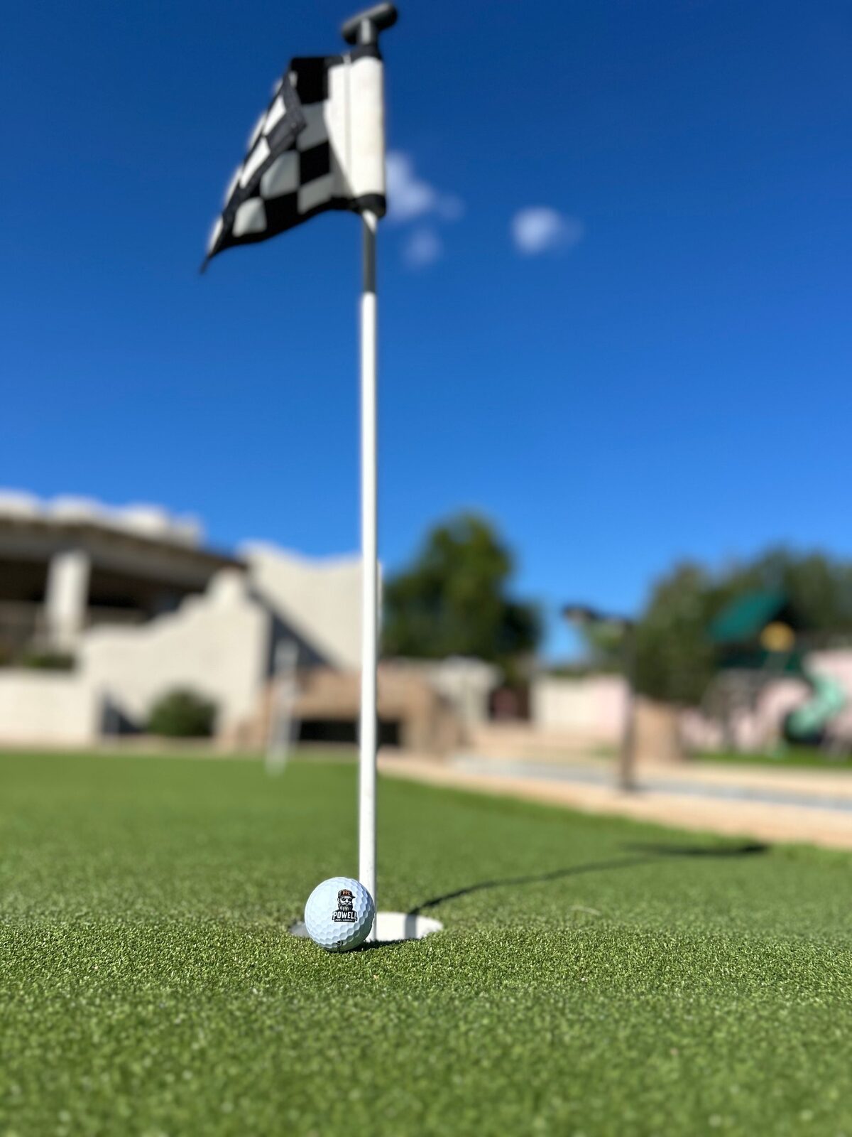 Close-up of a golf ball embossed with the Powell Pool Co logo, resting beside a cup on the putting green, checkered flag above