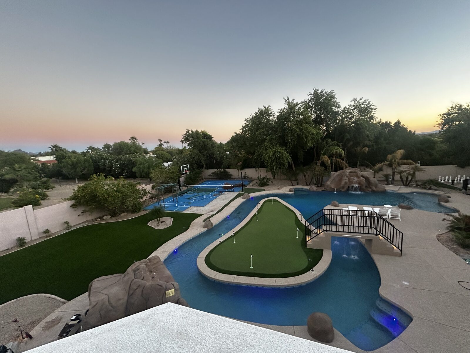 Elevated sunset view of the entire backyard — lazy river loop, lit putting-green island, bridge waterfall, rockslide grotto, and sport court