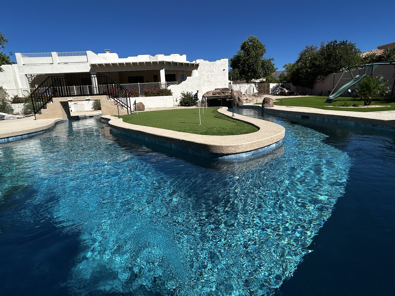 Water-level view across the vivid blue lazy river to the putting-green island, bridge, and the house beyond