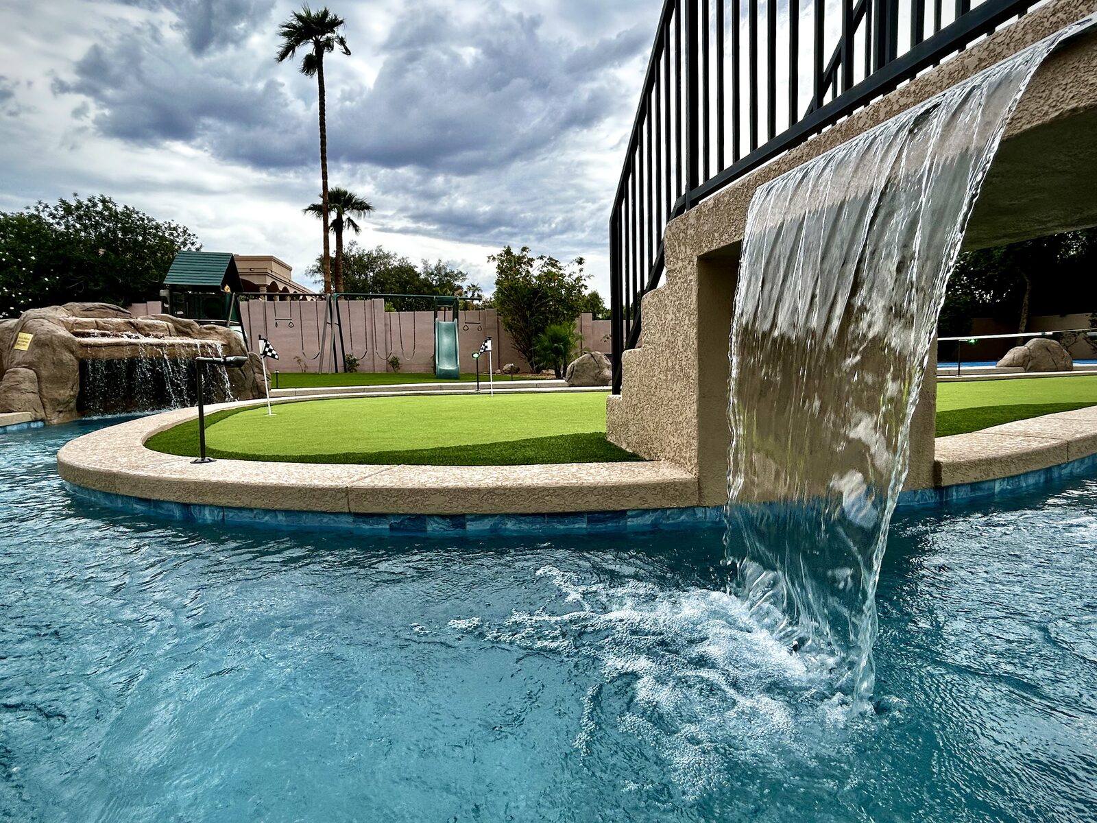 Close, low-angle view of the bridge's sheer-descent waterfall, with the putting green and swing set beyond