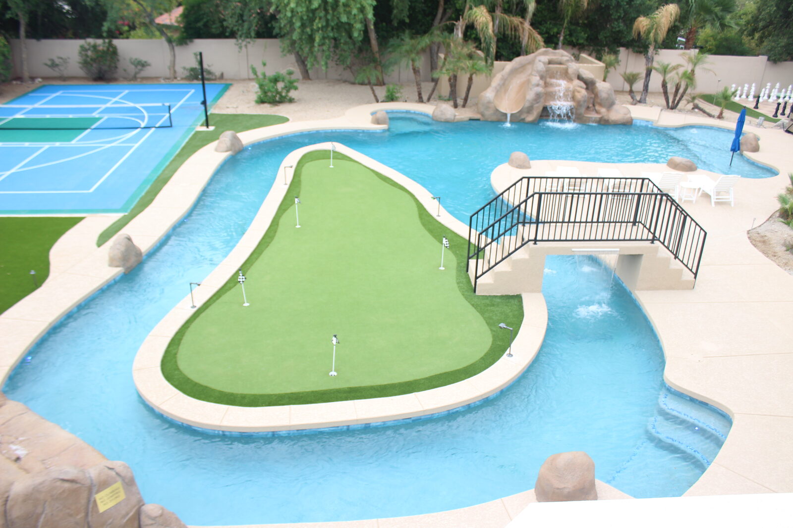 Elevated daytime view of the entire backyard — lazy river loop, putting-green island, bridge, rockslide grotto, sport court, and lounge area
