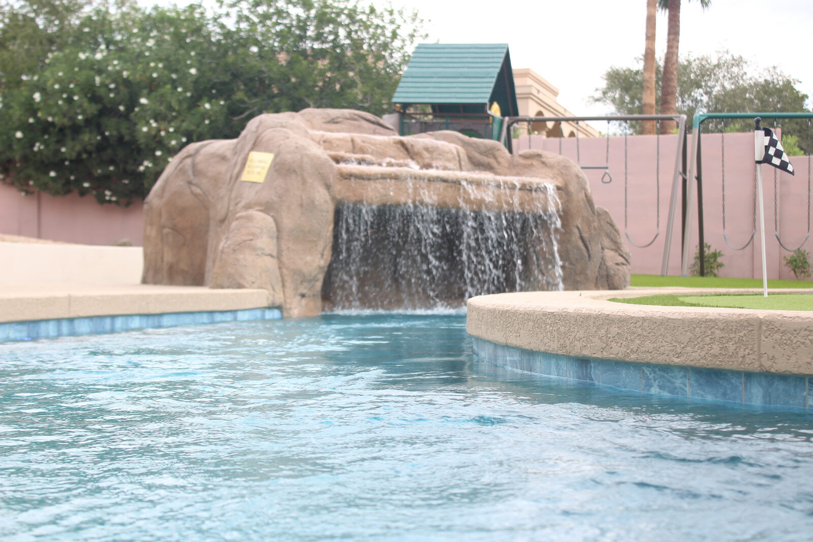 Water-level view across the lazy river toward the rockslide grotto waterfall, with the playground and putting green beyond