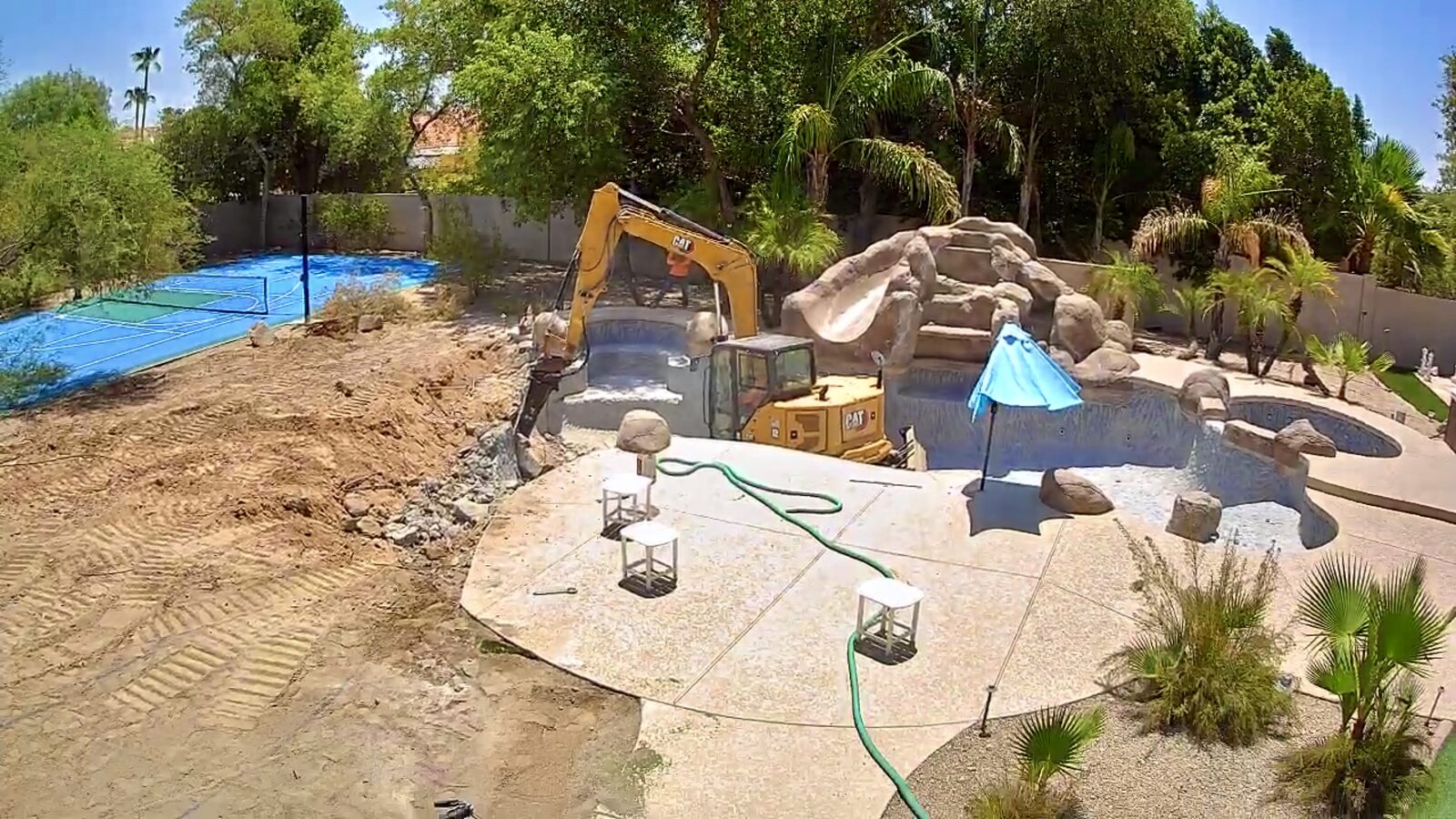 Ground-level view of the excavator mid-demolition, with the original grotto rockwork and tarped sport court behind it